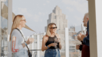 Three people holding drinks, chatting on a rooftop terrace with city buildings in the background on a sunny day.