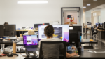 View from behind of an office worker at a desk with multiple computer screens, surrounded by a modern open office setup.