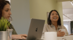 Two women in a relaxed office setting, one smiling with a cup in hand and the other focused on her laptop.