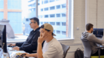 Three employees working at desks, concentrating on computer screens in an open-concept office with large windows.