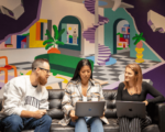 Three colleagues sitting on a couch with laptops, engaged in discussion. Behind them is a vibrant, abstract mural with bold colours and geometric shapes, giving a creative and modern atmosphere to the workspace.