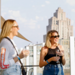 Two women holding drinks, chatting on a rooftop terrace with city buildings in the background on a sunny day.