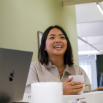 A woman in a relaxed office setting, smiling with a cup in hand. 