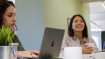 Two women in a relaxed office setting, one smiling with a cup in hand and the other focused on her laptop.